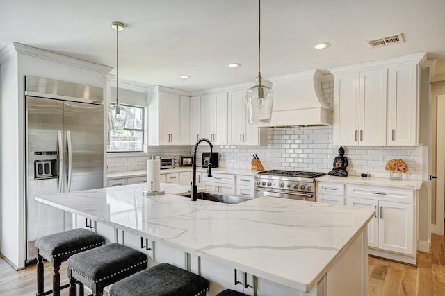 kitchen featuring premium range hood, stainless steel appliances, an island with sink, and decorative light fixtures