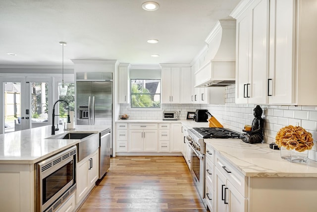 kitchen featuring light stone countertops, pendant lighting, white cabinets, built in appliances, and premium range hood