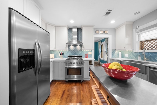 kitchen with hardwood / wood-style floors, appliances with stainless steel finishes, wall chimney range hood, pendant lighting, and white cabinets