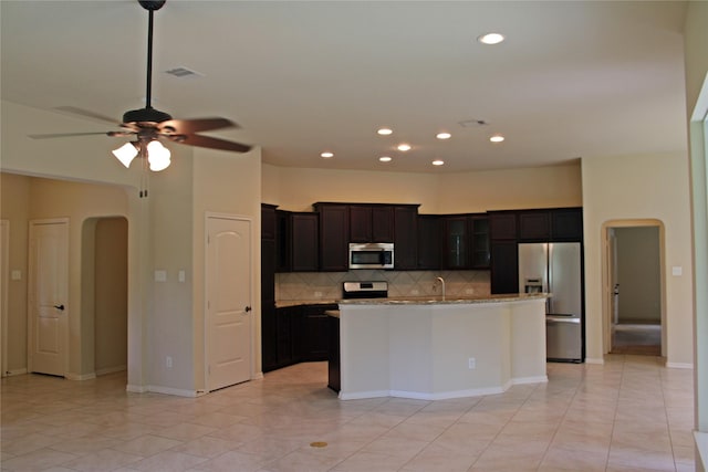 kitchen featuring a kitchen island with sink, stainless steel appliances, arched walkways, backsplash, and glass insert cabinets