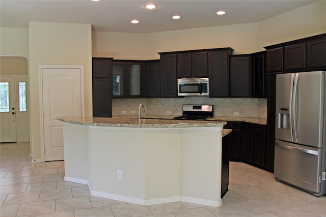 kitchen featuring an island with sink, stainless steel appliances, backsplash, light stone countertops, and glass insert cabinets