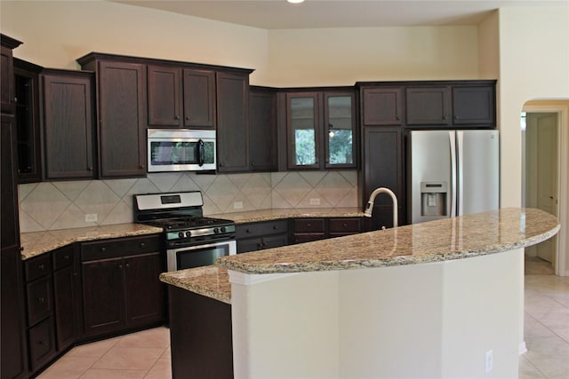 kitchen featuring appliances with stainless steel finishes, light tile patterned flooring, a center island with sink, and glass insert cabinets