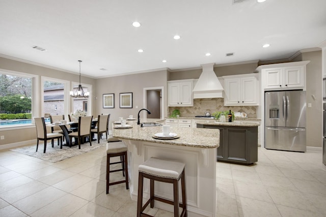 kitchen with white cabinetry, custom exhaust hood, an island with sink, stainless steel refrigerator with ice dispenser, and sink