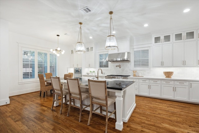 kitchen with dark wood-type flooring, decorative light fixtures, a center island with sink, tasteful backsplash, and white cabinetry