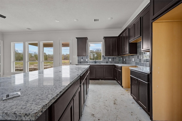 kitchen featuring decorative backsplash, dark brown cabinets, light stone countertops, a kitchen island, and crown molding