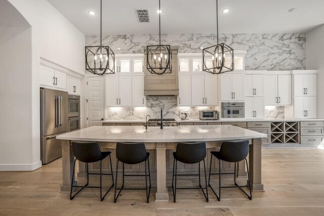 kitchen with appliances with stainless steel finishes, light wood-type flooring, a large island with sink, white cabinets, and decorative light fixtures