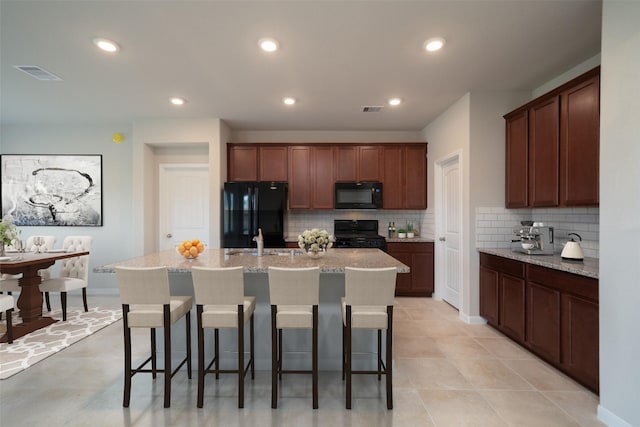 kitchen featuring sink, light stone counters, an island with sink, decorative backsplash, and black appliances