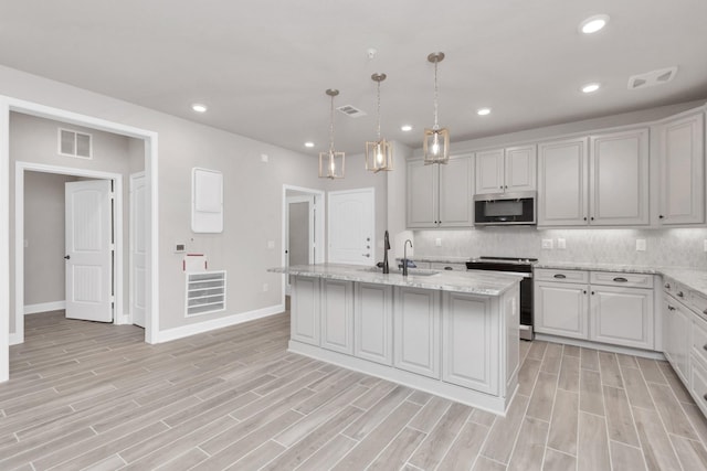 kitchen with a kitchen island with sink, appliances with stainless steel finishes, hanging light fixtures, light stone counters, and backsplash