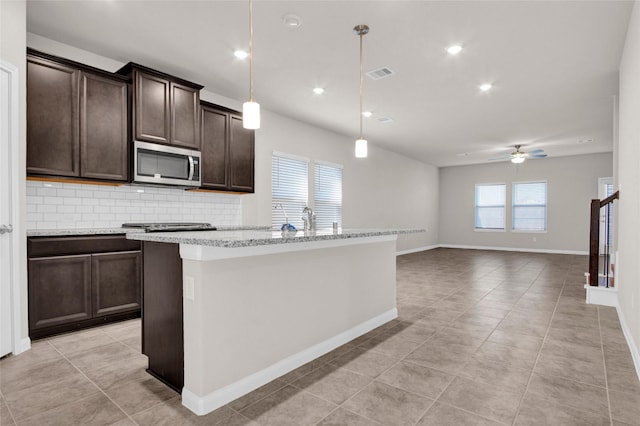 kitchen featuring decorative light fixtures, tasteful backsplash, a kitchen island with sink, ceiling fan, and dark brown cabinetry