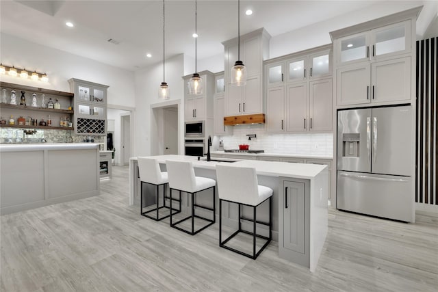 kitchen with decorative light fixtures, stainless steel appliances, an island with sink, tasteful backsplash, and light wood-type flooring