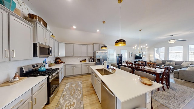 kitchen with a kitchen island with sink, stainless steel appliances, hanging light fixtures, sink, and ceiling fan with notable chandelier