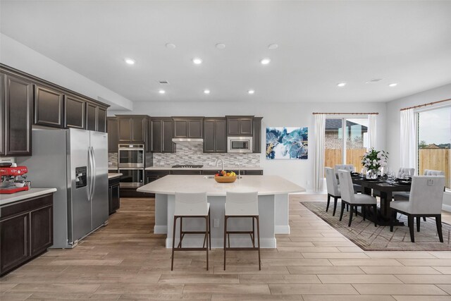 kitchen featuring a kitchen bar, tasteful backsplash, dark brown cabinets, a center island with sink, and appliances with stainless steel finishes