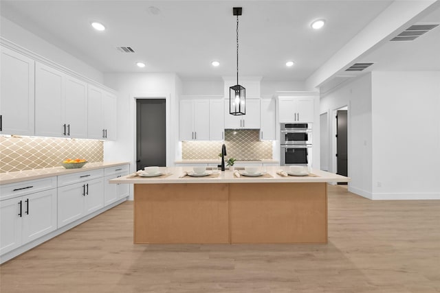 kitchen featuring decorative light fixtures, stainless steel double oven, a kitchen island with sink, and white cabinetry