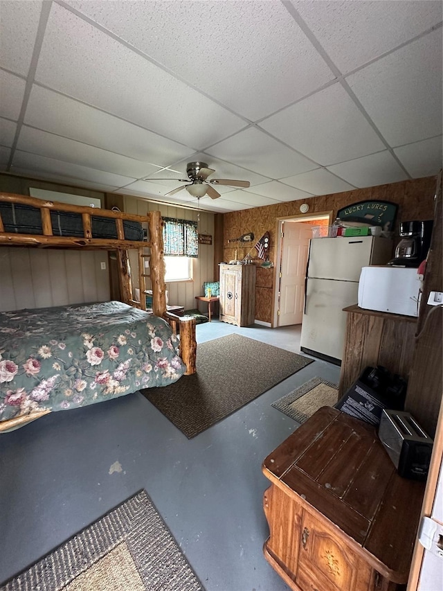 bedroom featuring white refrigerator, wooden walls, ceiling fan, a paneled ceiling, and concrete floors