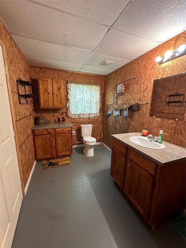 bathroom with wood walls, toilet, concrete floors, vanity, and a drop ceiling