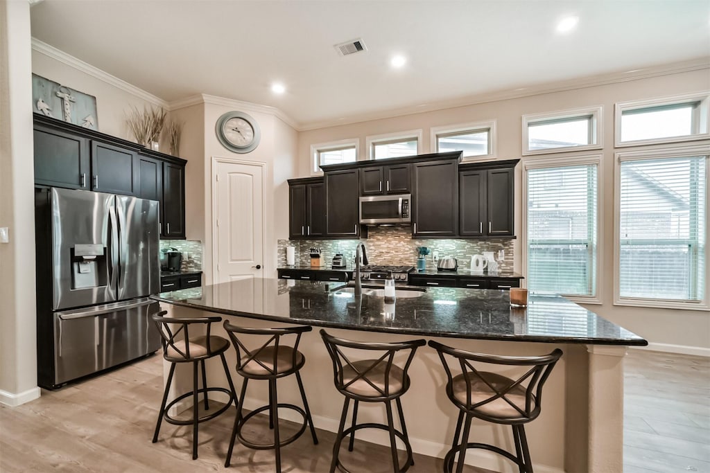 kitchen featuring crown molding, appliances with stainless steel finishes, an island with sink, dark stone counters, and decorative backsplash