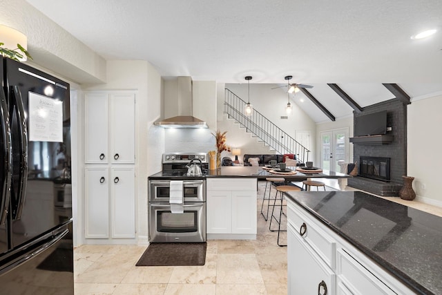 kitchen featuring range with two ovens, wall chimney range hood, black refrigerator, white cabinetry, and a fireplace