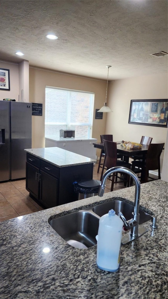 kitchen with light stone counters, a textured ceiling, pendant lighting, stainless steel refrigerator with ice dispenser, and sink