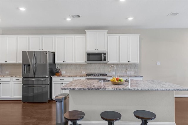 kitchen with stainless steel appliances, a kitchen island with sink, light stone countertops, and white cabinetry