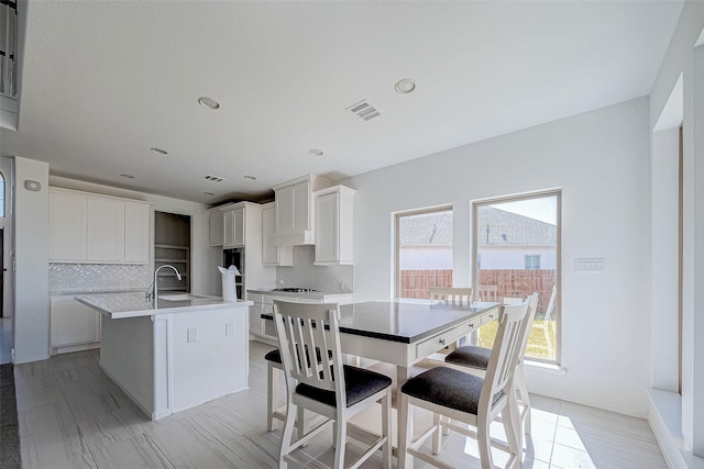 kitchen featuring a kitchen island with sink, light tile patterned floors, white cabinets, backsplash, and sink