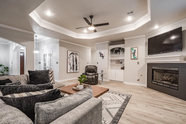 living room featuring a tile fireplace, ornamental molding, a tray ceiling, and light wood-type flooring