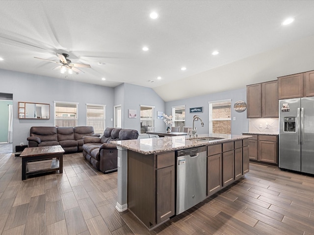 kitchen featuring an island with sink, lofted ceiling, sink, light stone counters, and stainless steel appliances