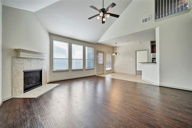 unfurnished living room featuring baseboard heating, a tile fireplace, light wood-type flooring, high vaulted ceiling, and ceiling fan with notable chandelier