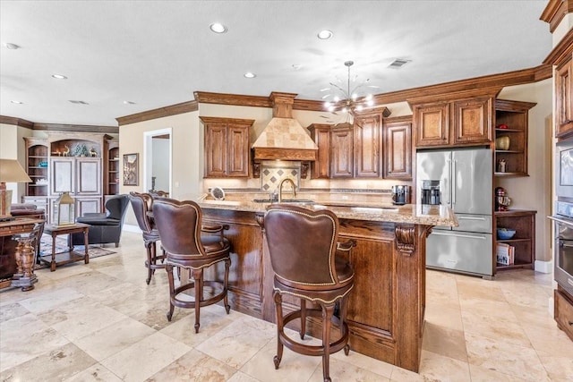 kitchen featuring stainless steel appliances, custom range hood, a large island with sink, light stone countertops, and crown molding