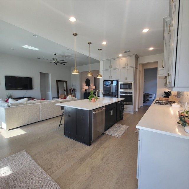 kitchen with white cabinets, light hardwood / wood-style flooring, decorative light fixtures, stainless steel appliances, and a center island with sink