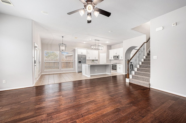 unfurnished living room featuring ceiling fan with notable chandelier, visible vents, baseboards, stairs, and light wood-type flooring