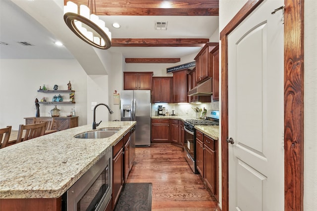 kitchen with light stone counters, stainless steel appliances, a center island with sink, beam ceiling, and sink