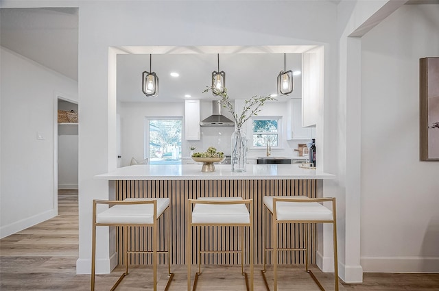 kitchen with a kitchen breakfast bar, pendant lighting, wall chimney exhaust hood, sink, and white cabinetry