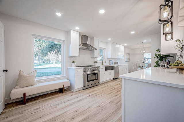 kitchen with white cabinets, light hardwood / wood-style flooring, wall chimney range hood, pendant lighting, and appliances with stainless steel finishes