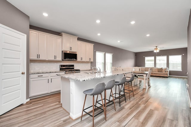 kitchen with light stone counters, ceiling fan, an island with sink, backsplash, and appliances with stainless steel finishes