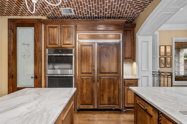 kitchen featuring double oven, brick ceiling, paneled built in fridge, light stone countertops, and light hardwood / wood-style flooring