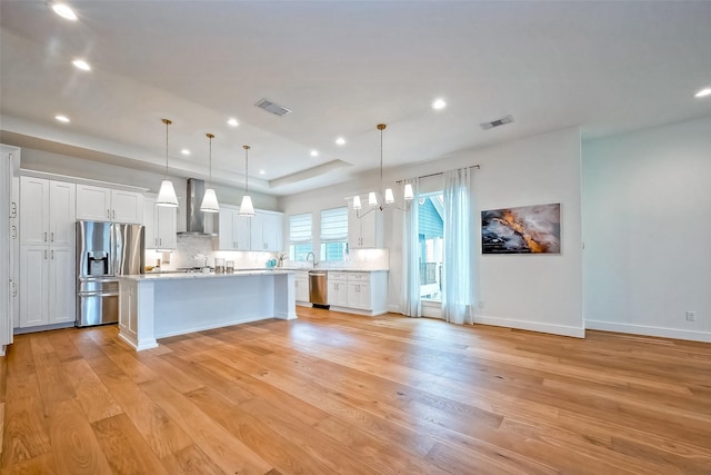 kitchen with decorative light fixtures, stainless steel appliances, wall chimney range hood, a kitchen island with sink, and white cabinets