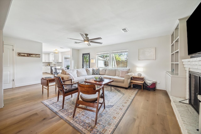 living room with a brick fireplace, built in shelves, light hardwood / wood-style flooring, and ceiling fan