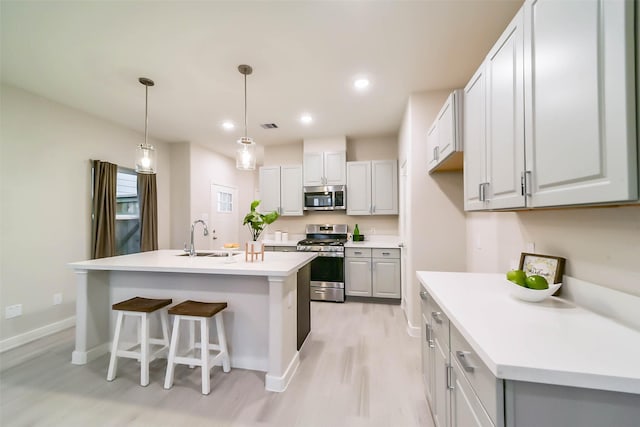 kitchen with appliances with stainless steel finishes, hanging light fixtures, an island with sink, sink, and light hardwood / wood-style flooring