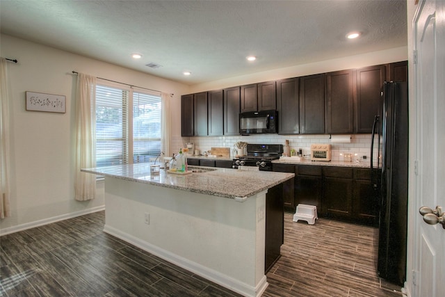 kitchen with light stone counters, black appliances, a kitchen island with sink, backsplash, and dark brown cabinets