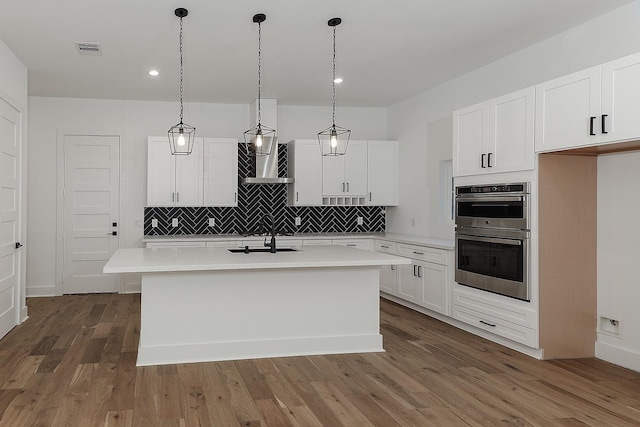 kitchen with a kitchen island with sink, double oven, dark hardwood / wood-style floors, white cabinetry, and wall chimney range hood