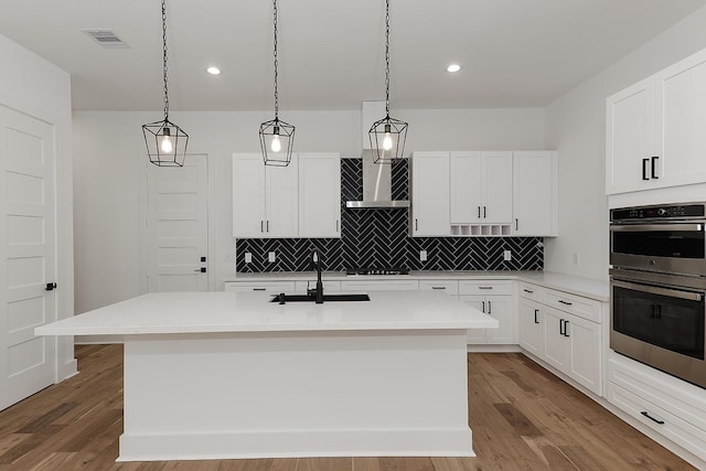 kitchen with sink, white cabinets, an island with sink, hanging light fixtures, and double oven