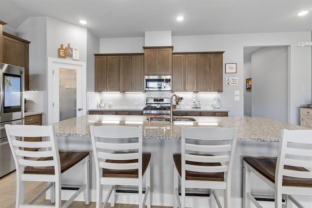 kitchen featuring stainless steel appliances, an island with sink, light stone countertops, a kitchen bar, and sink