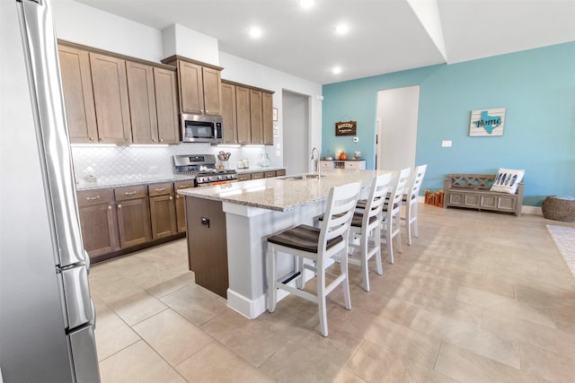 kitchen with light stone countertops, stainless steel appliances, an island with sink, a kitchen bar, and backsplash