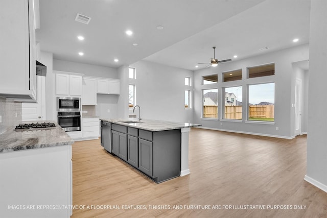 kitchen with white cabinets, ceiling fan, an island with sink, backsplash, and light stone countertops