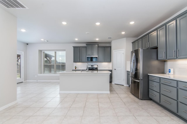 kitchen featuring stainless steel appliances, light tile patterned flooring, tasteful backsplash, an island with sink, and gray cabinetry