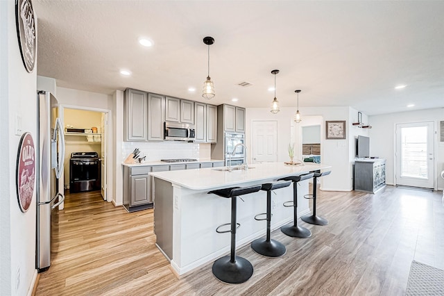 kitchen with stainless steel appliances, tasteful backsplash, light wood-type flooring, a kitchen island with sink, and gray cabinetry