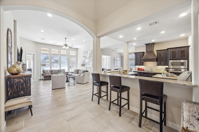 kitchen with ceiling fan, custom exhaust hood, a kitchen bar, light stone countertops, and stainless steel appliances