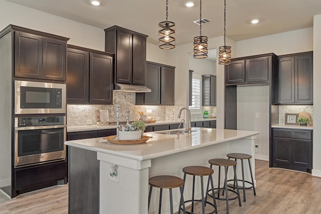 kitchen featuring an island with sink, sink, stainless steel oven, and decorative light fixtures