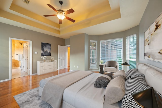 bedroom featuring ceiling fan, light wood-type flooring, ensuite bathroom, and a tray ceiling