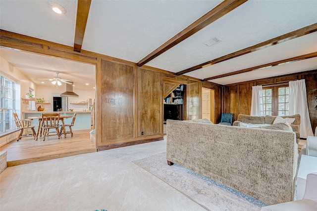 living room with a textured ceiling, wooden walls, light colored carpet, ceiling fan, and beamed ceiling